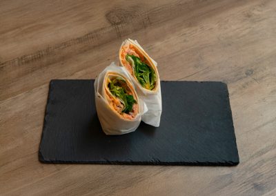 Der „Ma Owilli Ai Pai“ Lachs-Wrap von Bowls4 in Gera, halbiert und in Pergamentpapier gewickelt, serviert auf einer schwarzen Schieferplatte. Im Inneren sind Räucherlachs, Feldsalat und Mango deutlich sichtbar. Auch glutenfrei erhältlich.