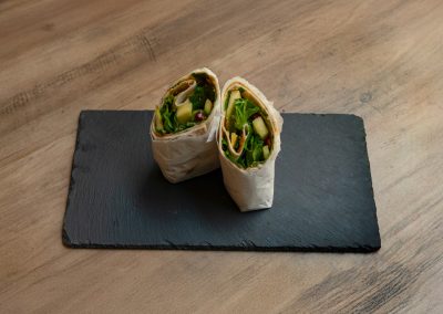 Der vegane „Ma Owilli Berena“ Wrap von Bowls4 in Gera, halbiert auf einer Schieferplatte. Im Querschnitt sind Guacamole, Mango-Stücke, Feldsalat, Granatapfelkerne und Sesam gut zu erkennen. Auch glutenfrei erhältlich