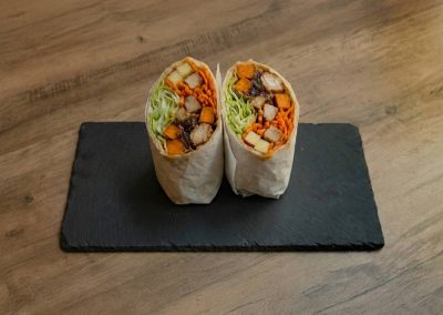 Nahaufnahme eines halbierten „Ma'Õwilli Inoa“ Wraps von Bowls4 in Gera, angerichtet auf einer schwarzen Schieferplatte. Die Füllung zeigt goldene Süßkartoffeln, geraspelte Möhren, frischen Eisbergsalat, Apfelstückchen und saftiges Teriyaki-Hähnchen.