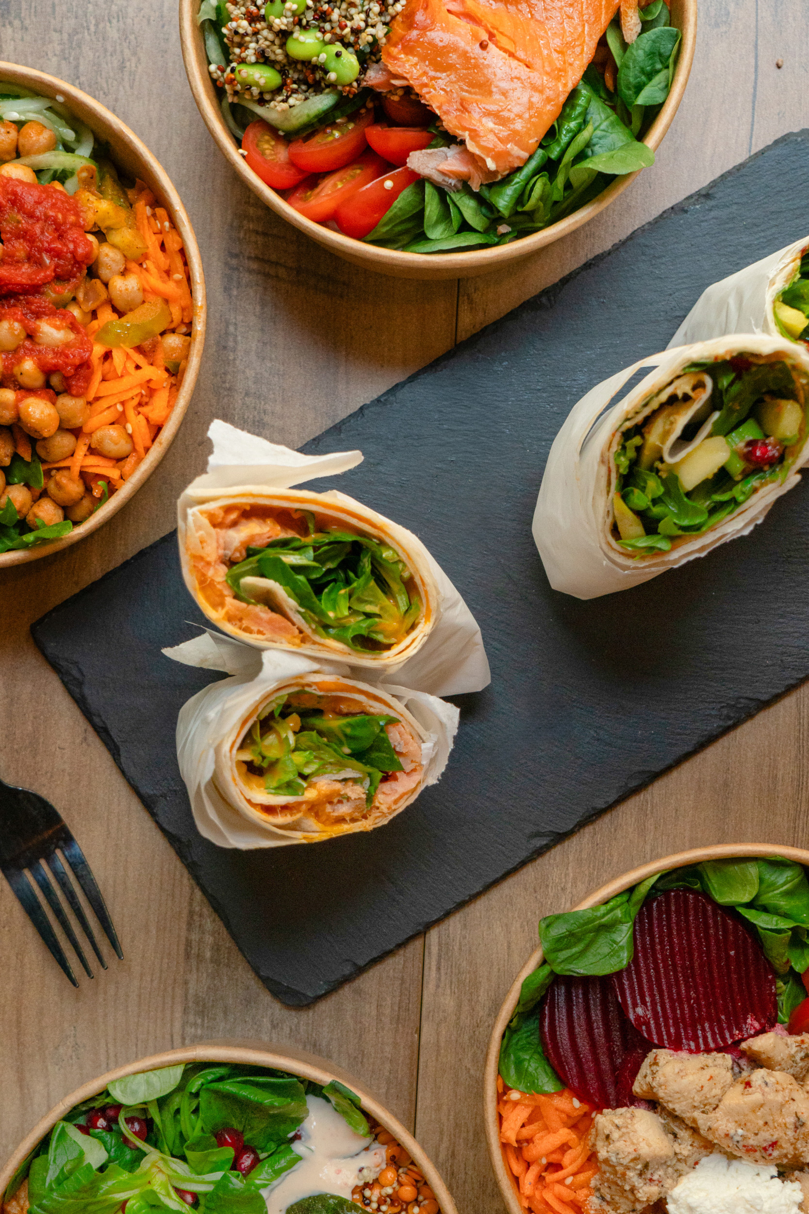 Eine bunte Auswahl an gesunden Gerichten von Bowls4 in Gera, fotografiert als Flatlay: In der Mitte ein aufgeschnittener Lachs-Wrap, umgeben von verschiedenen Bowls mit Lachs, Hähnchen, Roter Bete und Kichererbsen auf einem Holztisch.